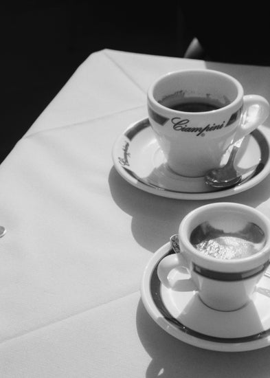 The Ciampini Poster captures a vintage Rome aesthetic with black and white photography of two coffee cups on saucers atop a white tablecloth, featuring contrasting sizes and shades with Ciampini elegantly inscribed, exuding timeless charm.