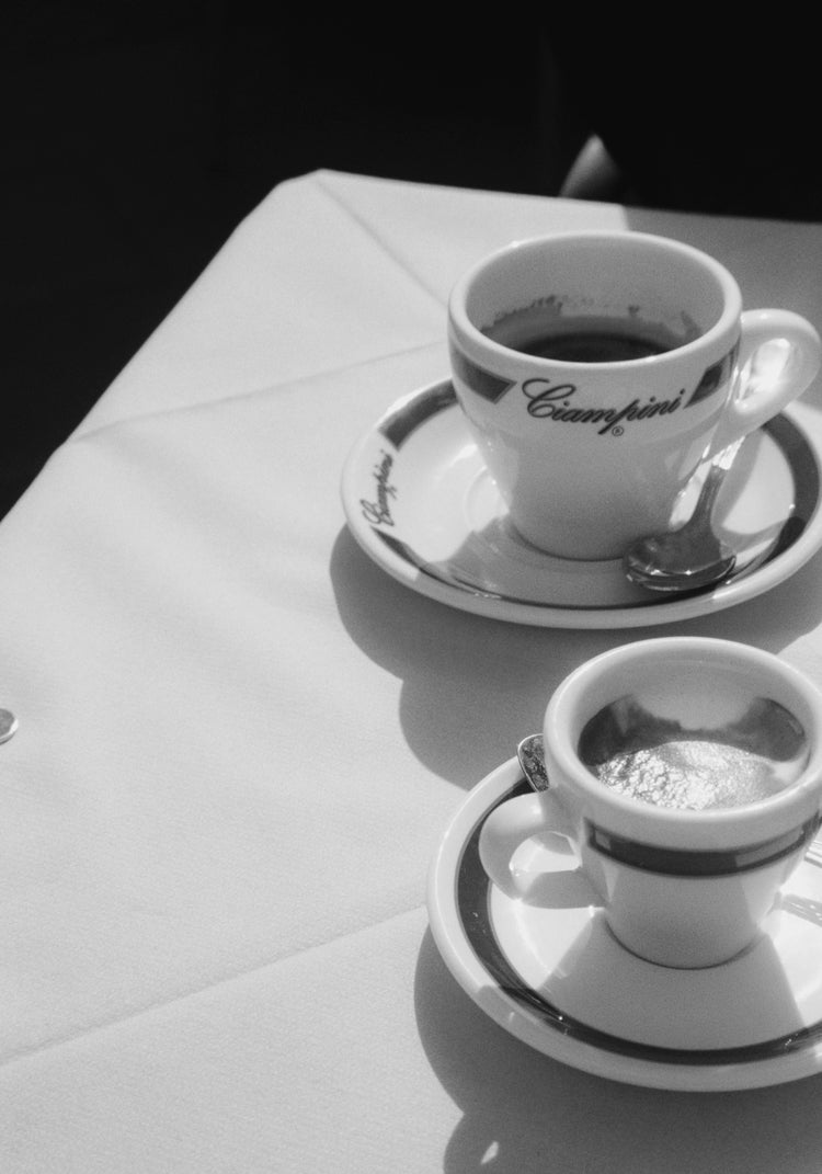 A black and white image showcases two espresso cups on saucers with spoons, set against gentle shadows on a white tablecloth. This scene evokes the vintage charm of Rome, reminiscent of a classic Ciampini Poster, with writing elegantly adorning the cups.