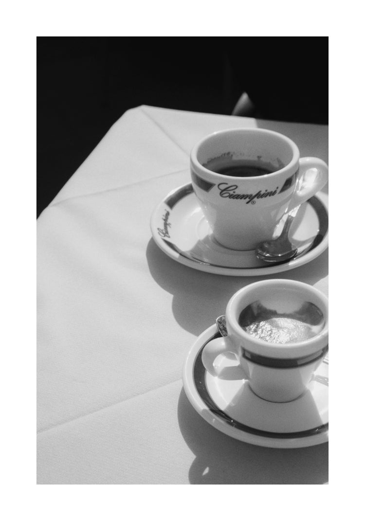 Amid a classic black and white Rome-inspired scene, two steaming coffee cups sit on a white tablecloth. The larger cup, bearing a logo and accompanied by a spoon on its saucer, is illuminated, reminiscent of an elegant Ciampini Poster.