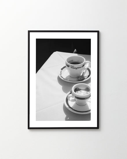 A black-and-white photo depicts two coffee cups on a table with a white tablecloth, evoking a classic Roman cafe. One cup holds espresso, while the larger cup labeled Cappuccino mirrors the classic charm of a Ciampini Poster.