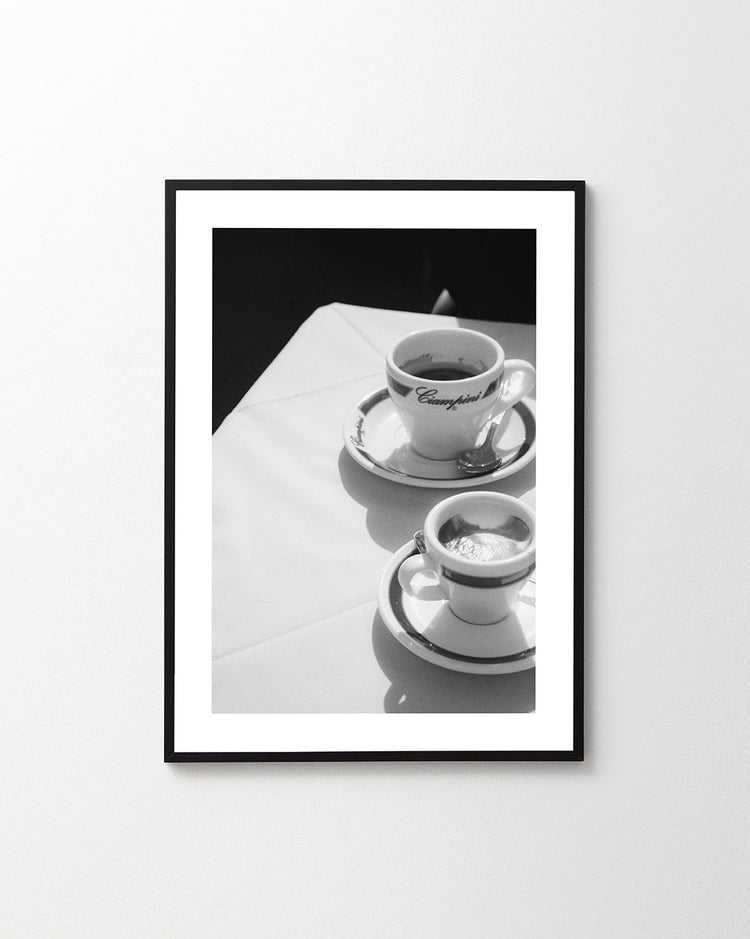 A black-and-white photo depicts two coffee cups on a table with a white tablecloth, evoking a classic Roman cafe. One cup holds espresso, while the larger cup labeled Cappuccino mirrors the classic charm of a Ciampini Poster.
