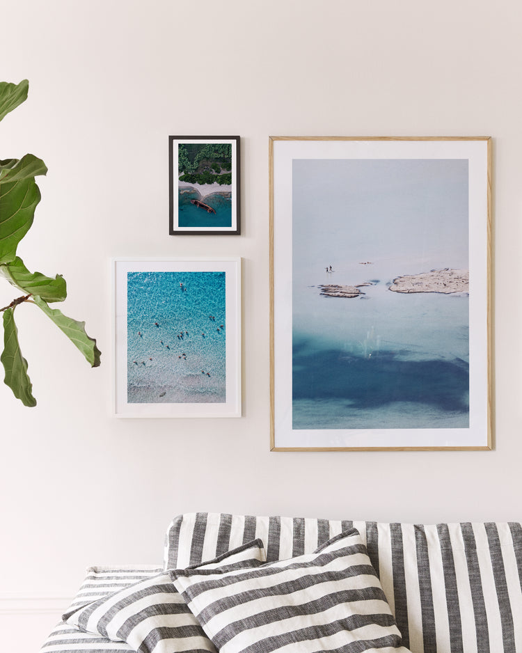 A beige wall showcases three framed photos: the Eavesdropping IX Poster depicting a beach scene with swimmers, another showing a rocky island in turquoise water, and a smaller serene beach view. A striped couch and leafy plant create an inviting space perfect for relaxation after a long day.