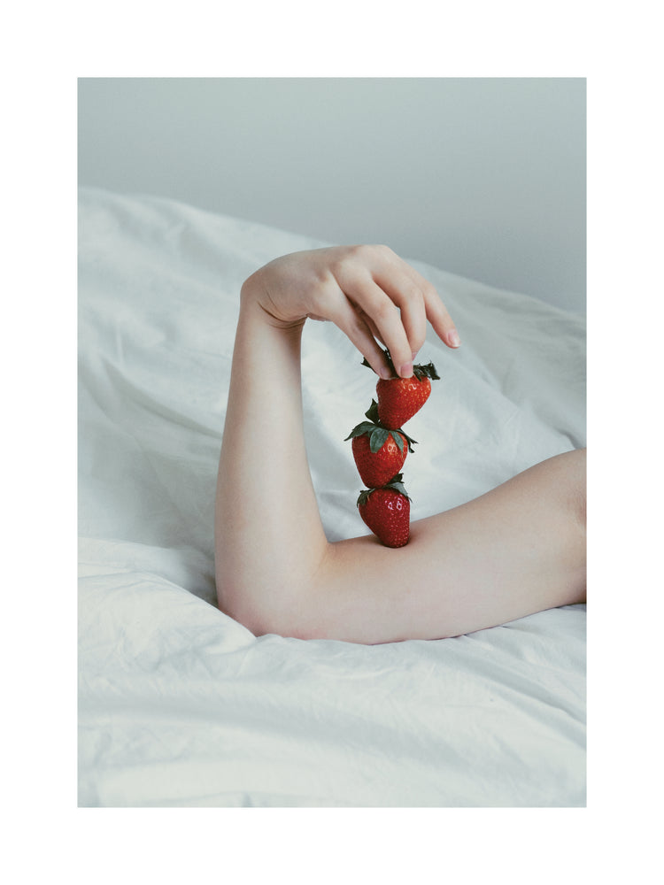 Female arm resting on a sheet with three strawberries stacked on her skin.
