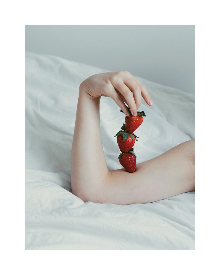 Female arm resting on a sheet with three strawberries stacked on her skin.
