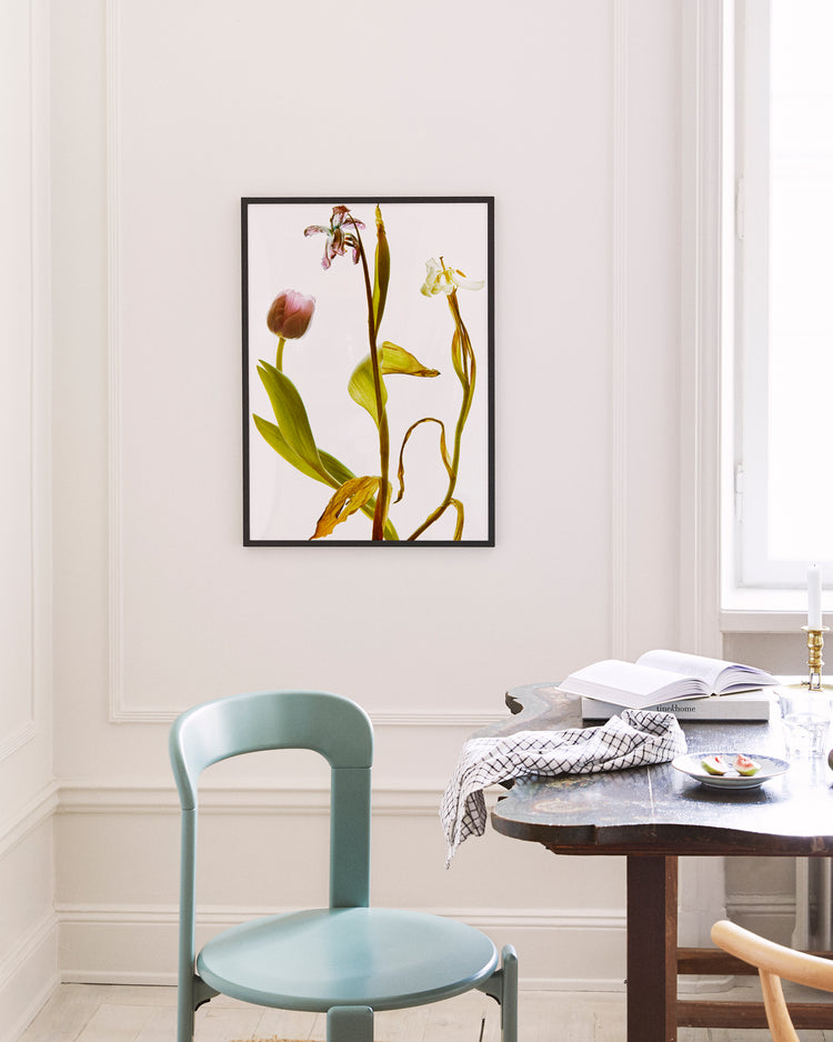A minimalist room features a light blue chair and wooden table. An open book and checkered cloth are placed on the table, while a framed Generations Poster showcasing flowers and leaves decorates the white wall.