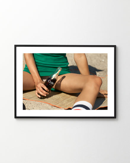 Dressed in green and relaxing on a mat outdoors, a person holds a Coca-Cola bottle, capturing the retro vibes of the Girls Just Want To Chill Out Poster. The partial view and sunny backdrop enhance its wall art appeal.