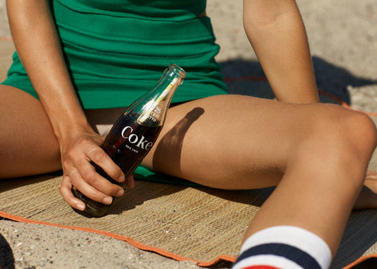 Woman in a green dress sitting on a beach and holding a glass bottle of coke.