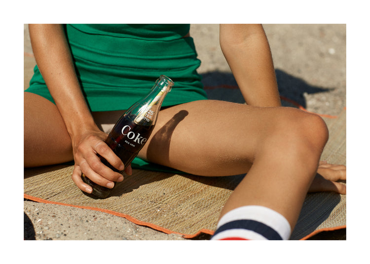 Woman in a green dress sitting on a beach and holding a glass bottle of coke.