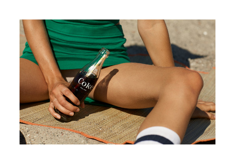 Woman in a green dress sitting on a beach and holding a glass bottle of coke.