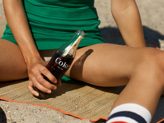 Woman in a green dress sitting on a beach and holding a glass bottle of coke.