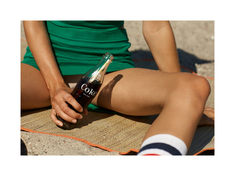 Woman in a green dress sitting on a beach and holding a glass bottle of coke.