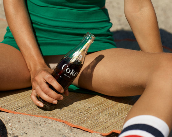 Woman in a green dress sitting on a beach and holding a glass bottle of coke.