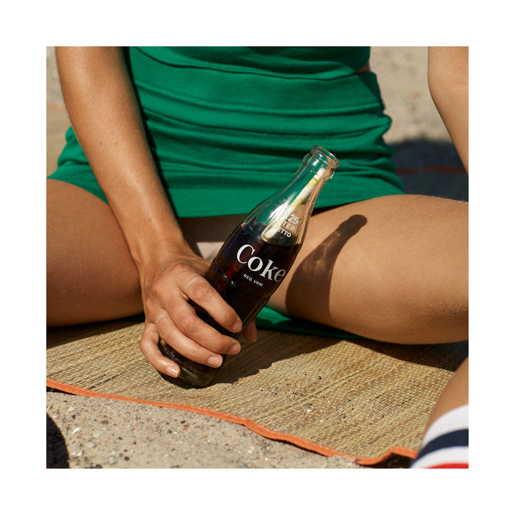 Woman in a green dress sitting on a beach and holding a glass bottle of coke.