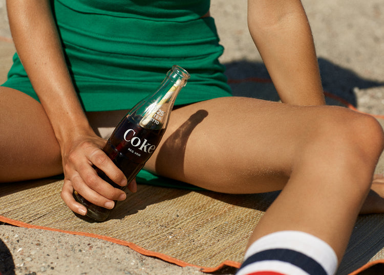 Woman in a green dress sitting on a beach and holding a glass bottle of coke.