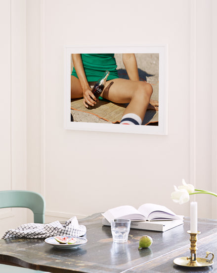 A framed Girls Just Want To Chill Out Poster on the white wall shows a person in green with a bottle on a mat. Below, the table holds an open book, cloth, water glass, candle, vase with white tulip; nearby is a partially visible green chair.
