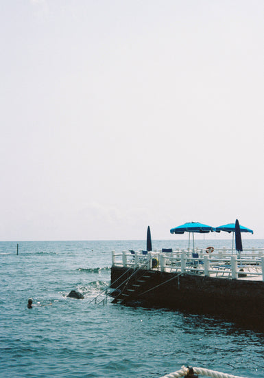 The Santa Marinella Poster portrays Italys sun-drenched coast with a serene scene of blue umbrellas and sunbathing chairs on a concrete platform. A ladder descends into the calm sea, where a swimmer enjoys the tranquil waters under a clear sky, crafting a romantic Mediterranean playground.