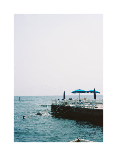The Santa Marinella Poster depicts a serene seaside platform with blue umbrellas and white railings extending over calm ocean waters on Italys sunny coast, capturing a person swimming nearby under clear skies for a tranquil, vacation-like ambiance.