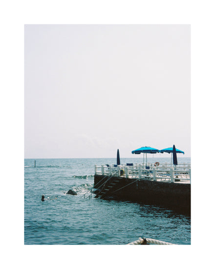 The Santa Marinella Poster features Italys sun-drenched coast with two blue umbrellas on the pier. Steps descend to the ocean, where someone swims by a clear sky and calm sea, capturing a perfect Mediterranean seascape.