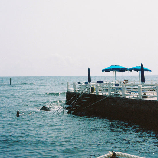 The Santa Marinella Poster captures a Mediterranean seascape with a concrete platform over the water, blue umbrellas, and sun loungers perfectly aligned. Steps descend to the serene sea, offering a romantic Italian swimming experience under sunny skies.
