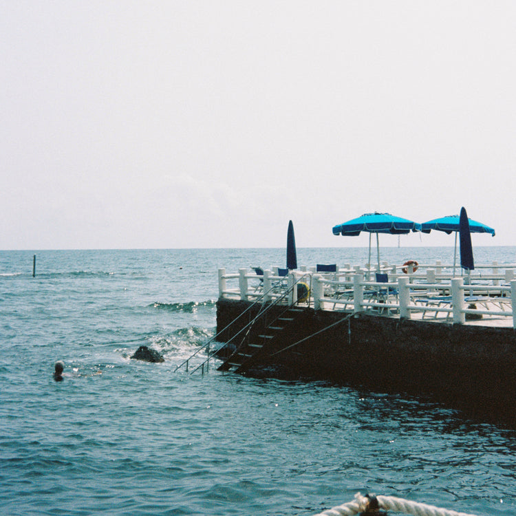 The Santa Marinella Poster captures a Mediterranean seascape with a concrete platform over the water, blue umbrellas, and sun loungers perfectly aligned. Steps descend to the serene sea, offering a romantic Italian swimming experience under sunny skies.