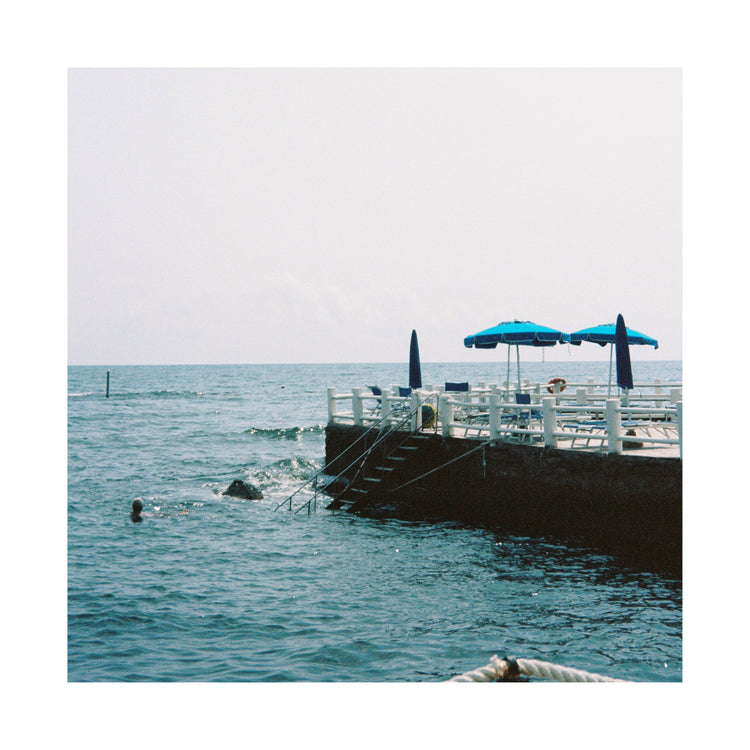 A Santa Marinella Poster depicts a serene Mediterranean scene: a wooden deck stretches into Italys sunny coast, with two blue umbrellas providing shade. A swimmer enjoys the calm, gentle waves of the ocean under a clear sky, showcasing an idyllic seaside landscape.