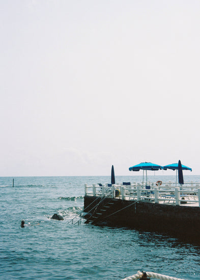 The Santa Marinella Poster depicts a tranquil Italian coastal scene with a pier over the water. A staircase leads down as blue umbrellas dot the landscape and a swimmer enjoys the serene setting, all under a clear, sunlit sky that evokes romance and tranquility.