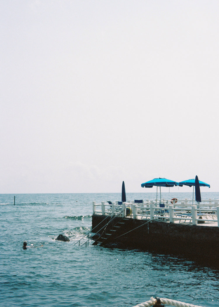 The Santa Marinella Poster depicts a tranquil Italian coastal scene with a pier over the water. A staircase leads down as blue umbrellas dot the landscape and a swimmer enjoys the serene setting, all under a clear, sunlit sky that evokes romance and tranquility.