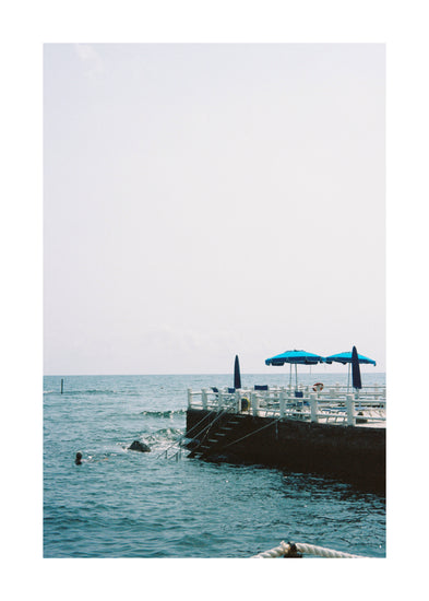 The Santa Marinella Poster depicts Italys sunny coast with a dock, steps into the water, and blue umbrellas on the deck. A swimmer enjoys the romantic sea playground beneath a clear sky.