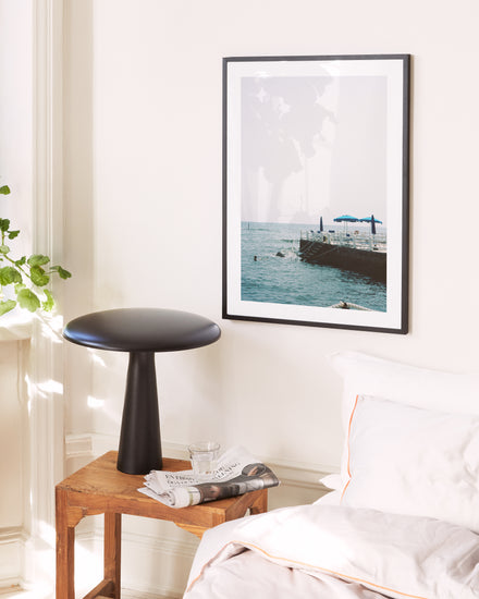 A serene bedroom captures Italys sunny coast, featuring a wooden table with a sleek black lamp, a glass, and a folded newspaper. A Santa Marinella Poster graces the wall near the white bed and lush green plant.