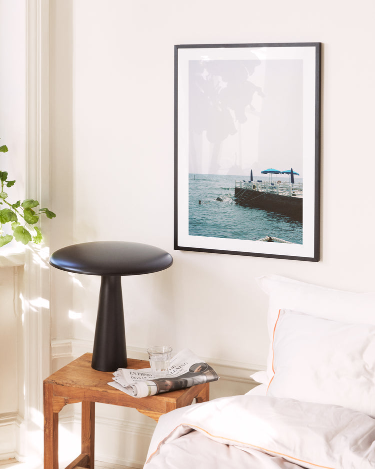 A serene bedroom captures Italys sunny coast, featuring a wooden table with a sleek black lamp, a glass, and a folded newspaper. A Santa Marinella Poster graces the wall near the white bed and lush green plant.