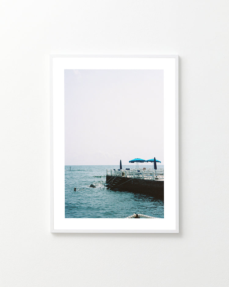 The Santa Marinella Poster depicts a tranquil Italian-inspired ocean scene with a concrete pier adorned with blue umbrellas. A swimmer enjoys the clear, sunny atmosphere reminiscent of Italys romantic coast.