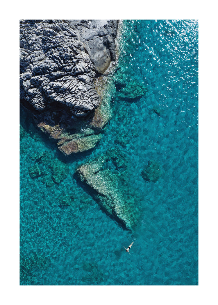 A small woman swimming alone in a big blue sea next to a grey cliff.