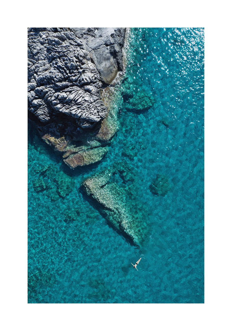 A small woman swimming alone in a big blue sea next to a grey cliff.