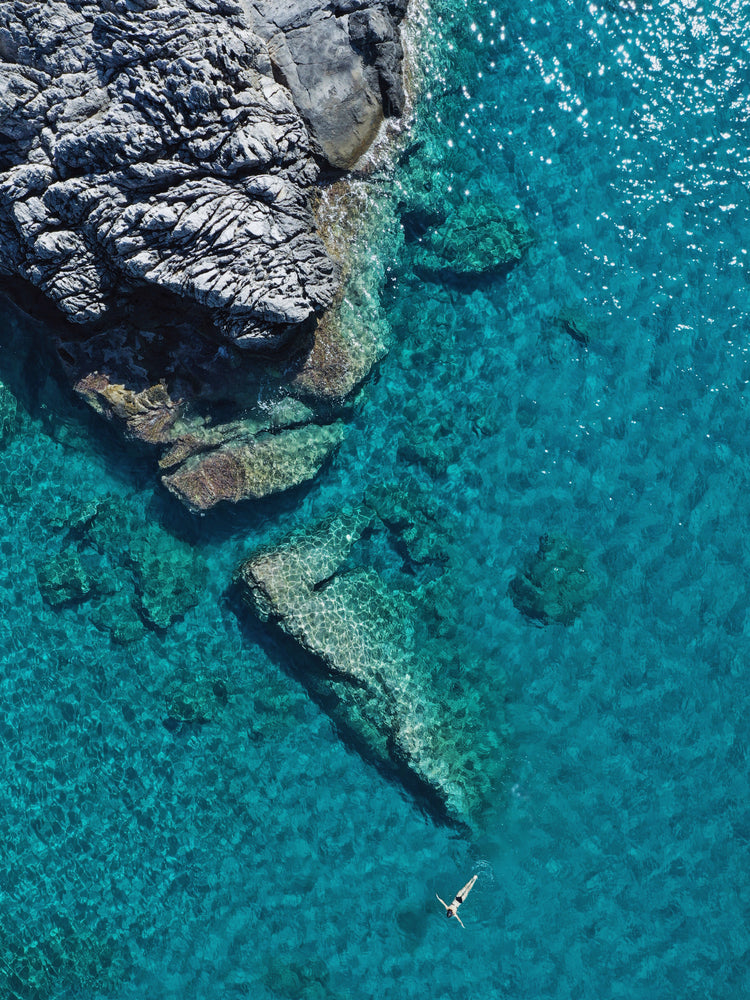 A small woman swimming alone in a big blue sea next to a grey cliff.