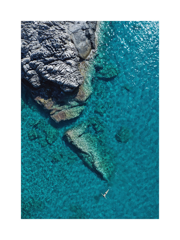 A small woman swimming alone in a big blue sea next to a grey cliff.