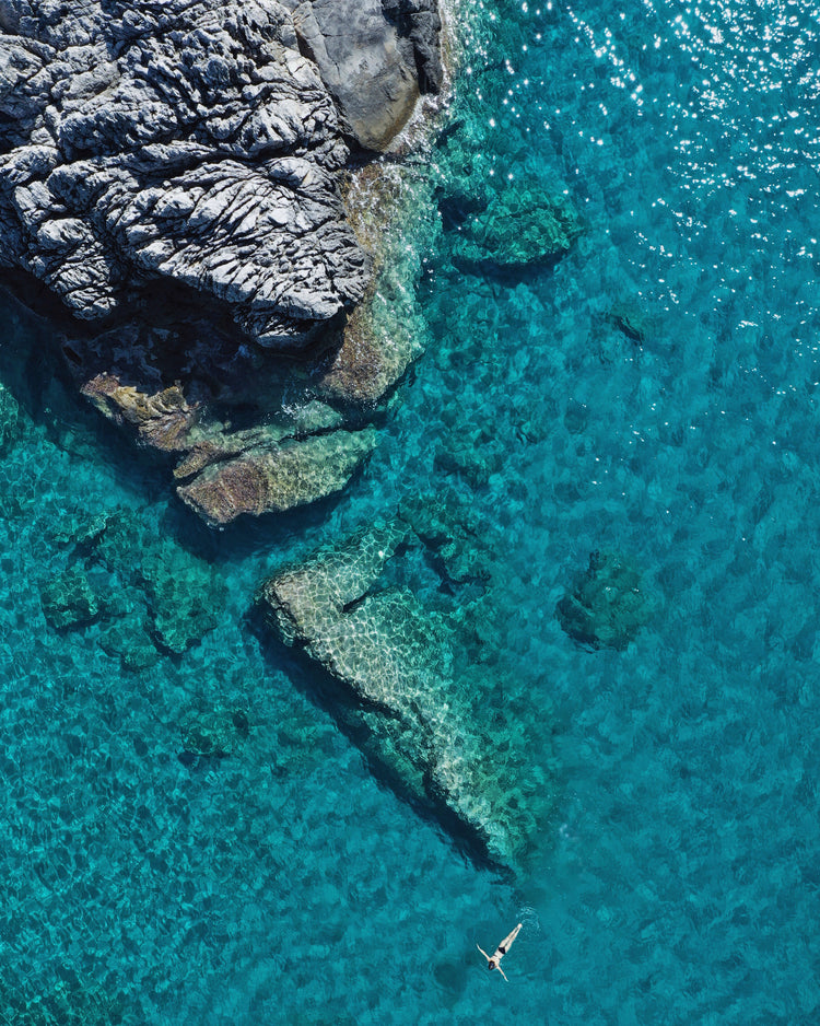 A small woman swimming alone in a big blue sea next to a grey cliff.