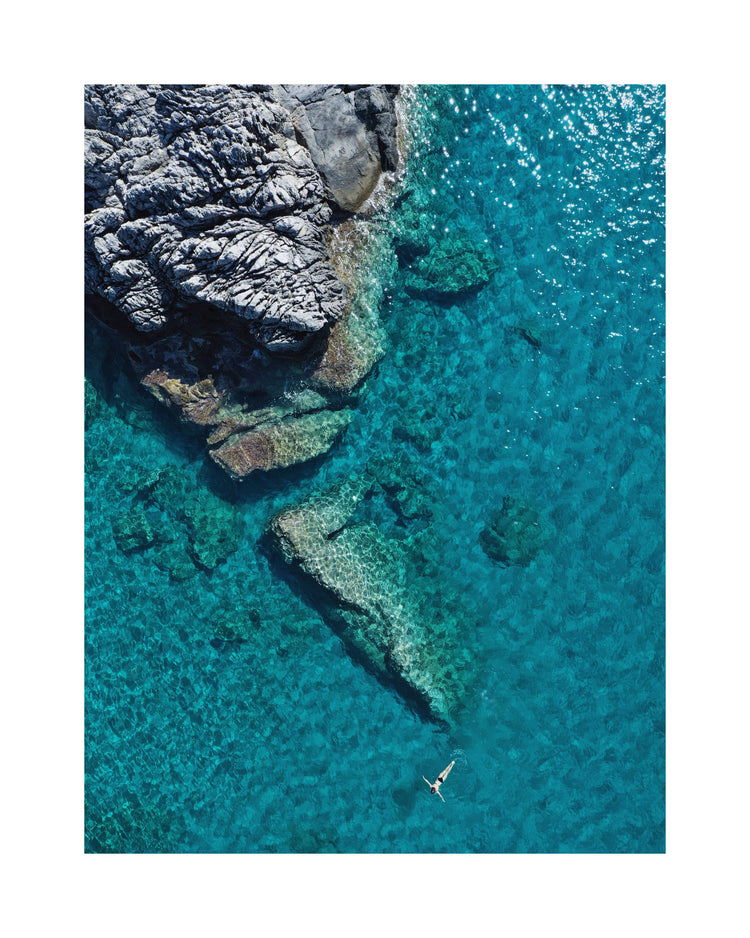 A small woman swimming alone in a big blue sea next to a grey cliff.