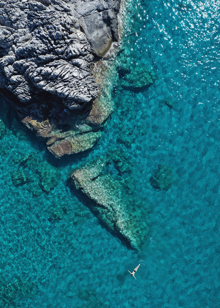 A small woman swimming alone in a big blue sea next to a grey cliff.