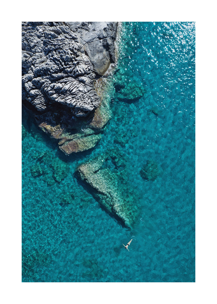 A small woman swimming alone in a big blue sea next to a grey cliff.