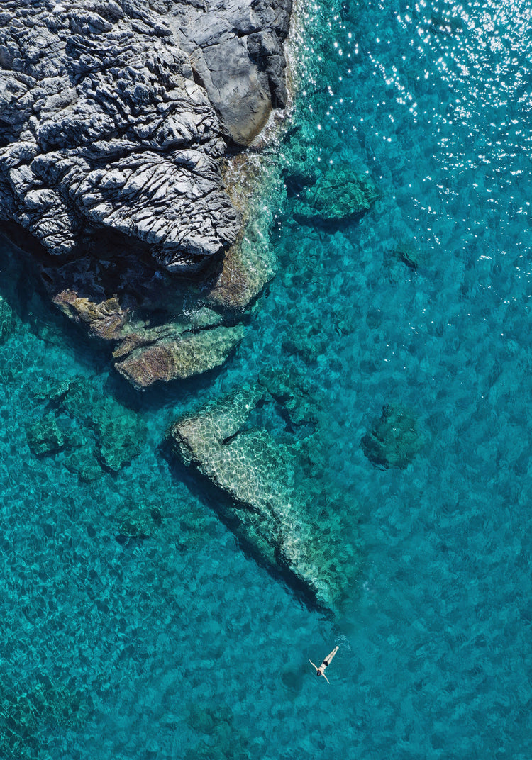 A small woman swimming alone in a big blue sea next to a grey cliff.