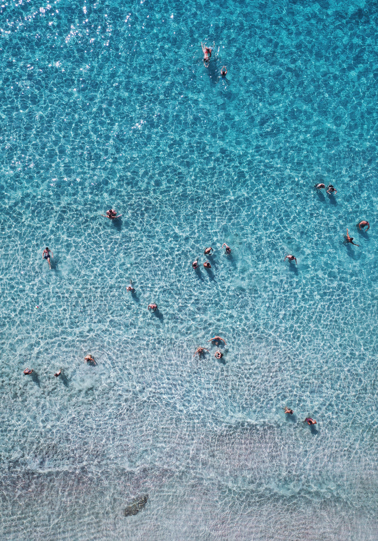 People swimming in the shallows in a blue, calm ocean close to a white beach.