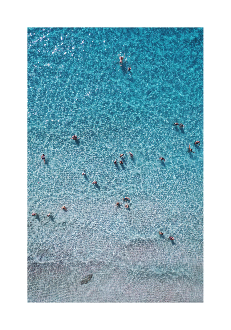 People swimming in the shallows in a blue, calm ocean close to a white beach.
