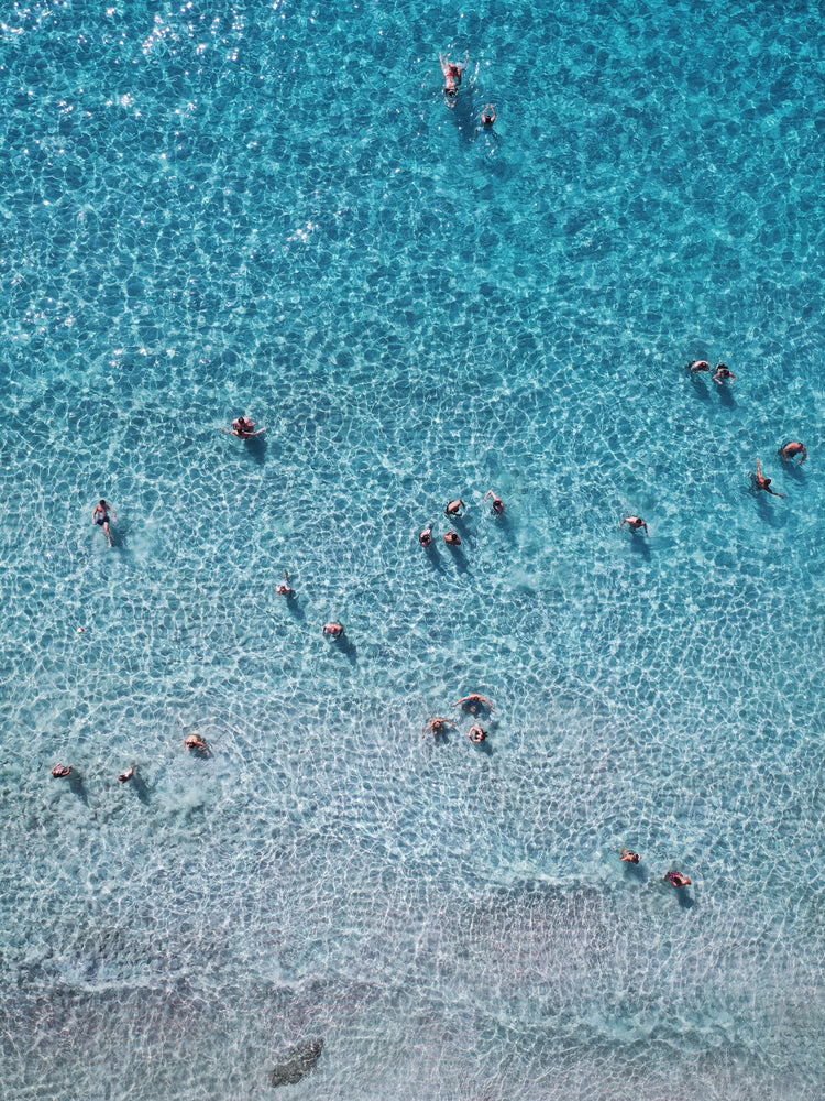 People swimming in the shallows in a blue, calm ocean close to a white beach.