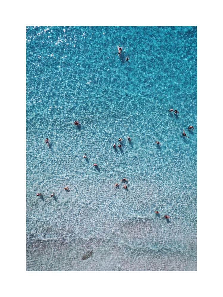People swimming in the shallows in a blue, calm ocean close to a white beach.