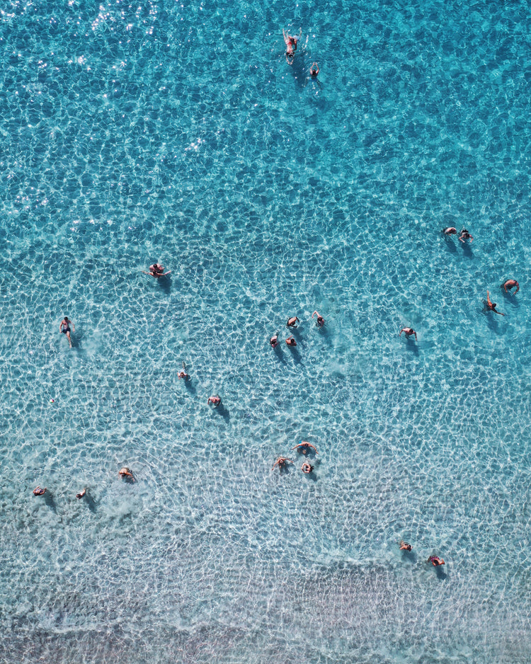 People swimming in the shallows in a blue, calm ocean close to a white beach.