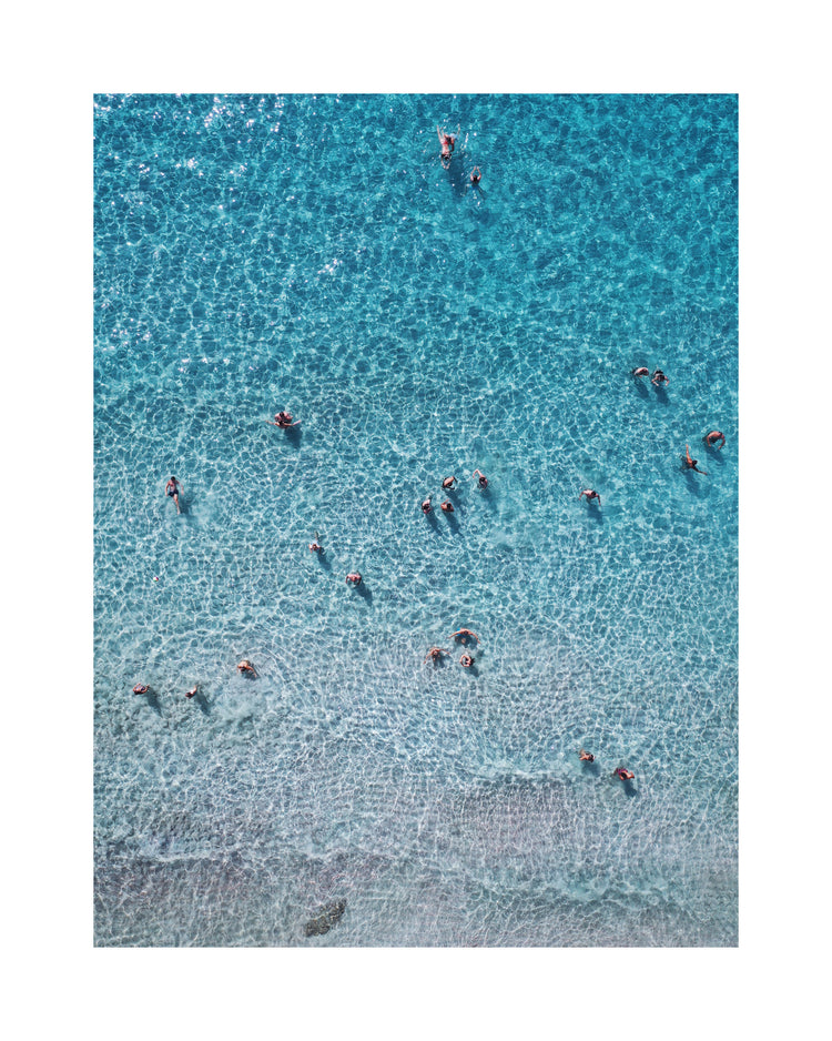 People swimming in the shallows in a blue, calm ocean close to a white beach.