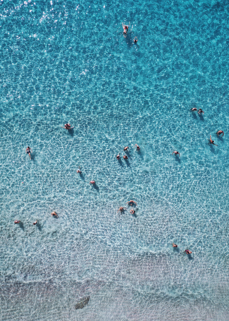 People swimming in the shallows in a blue, calm ocean close to a white beach.
