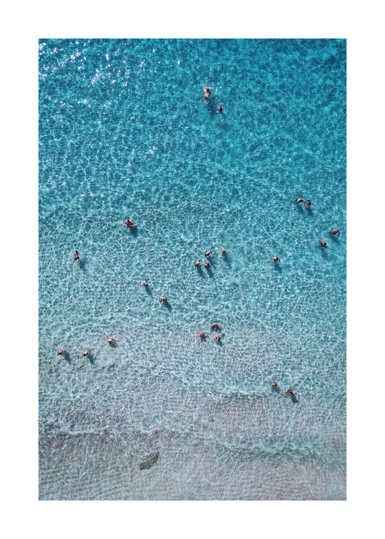 People swimming in the shallows in a blue, calm ocean close to a white beach.