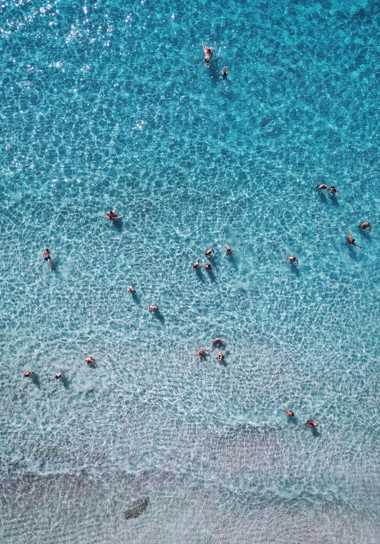 People swimming in the shallows in a blue, calm ocean close to a white beach.