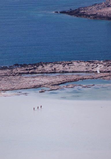 Three people walking in the shallows, heading towards the deep blue ocean ahead.